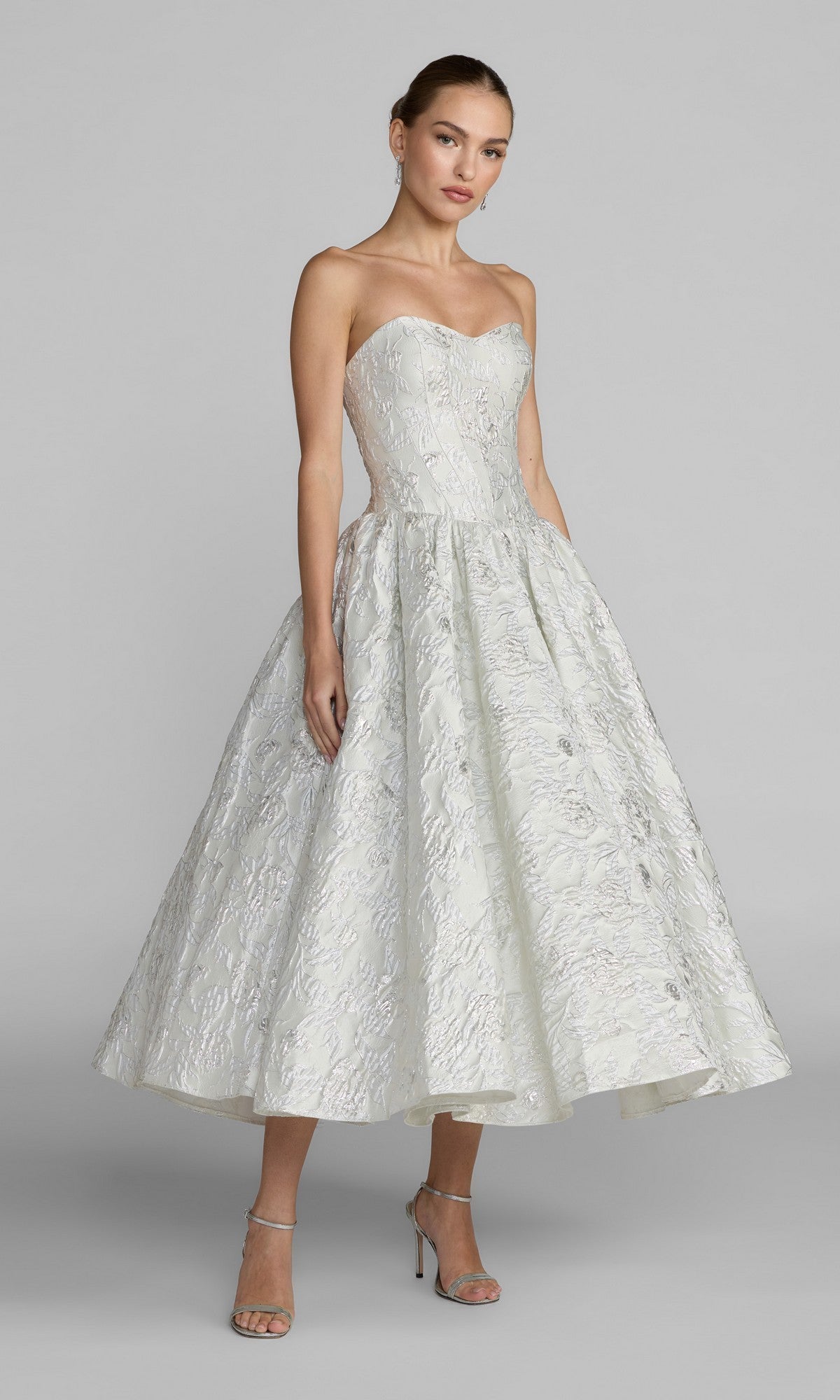 An elegant woman stands against a plain gray background wearing the ivory Tea-Length Party Dress: Mac Duggal 69146, featuring a strapless sweetheart bodice, textured midi A-line skirt with pockets, and paired with silver high-heeled sandals.